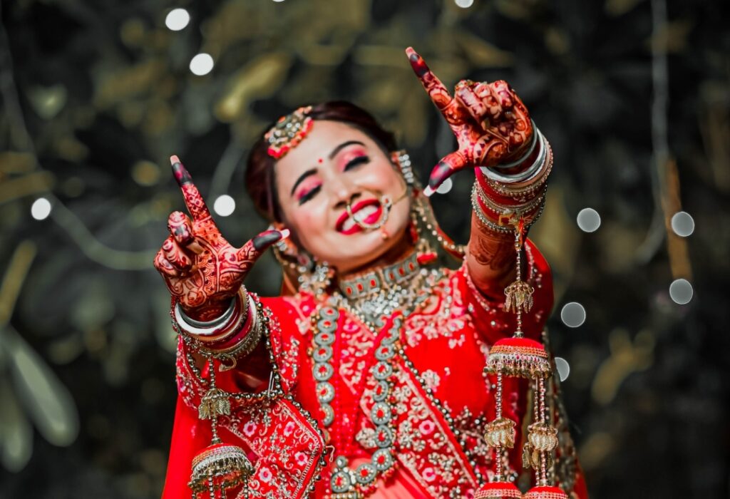 Indian bride smiling and showing intricate mehendi, wearing traditional red wedding attire, jewelry, and makeup.